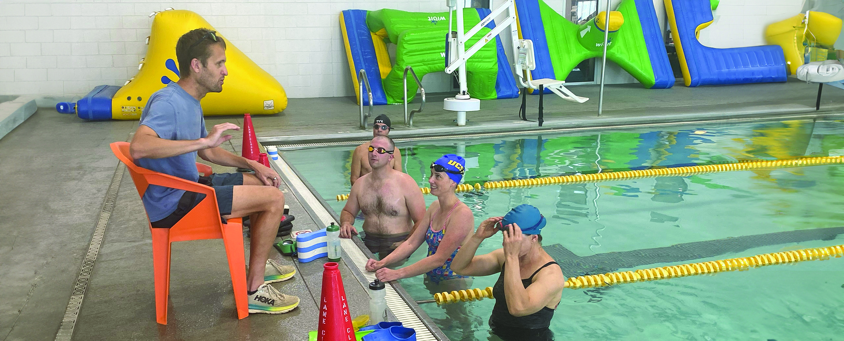 A masters swim coach seated beside an indoor pool during a training activity.