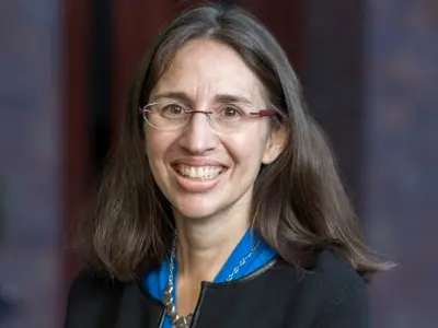 Headshot of a woman wearing glasses and a dark suit against a grey background.