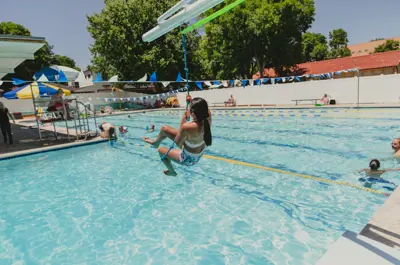 girl on a zipline at an outdoor pool near shaded deck seating.