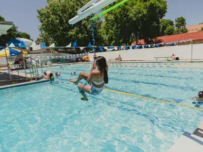 girl on a zipline at an outdoor pool near shaded deck seating.