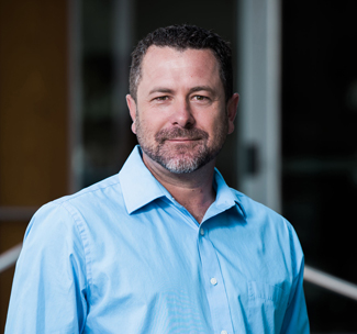 Headshot of a man wearing a blue shirt in front of a dark background.