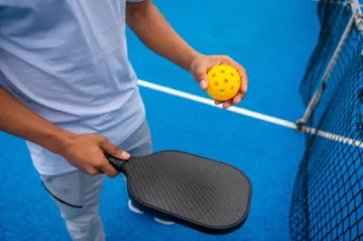 Player holding a paddle and ball beside a pickleball net.