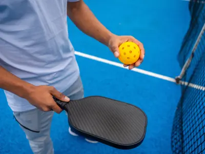 Player holding a paddle and ball beside a pickleball net.